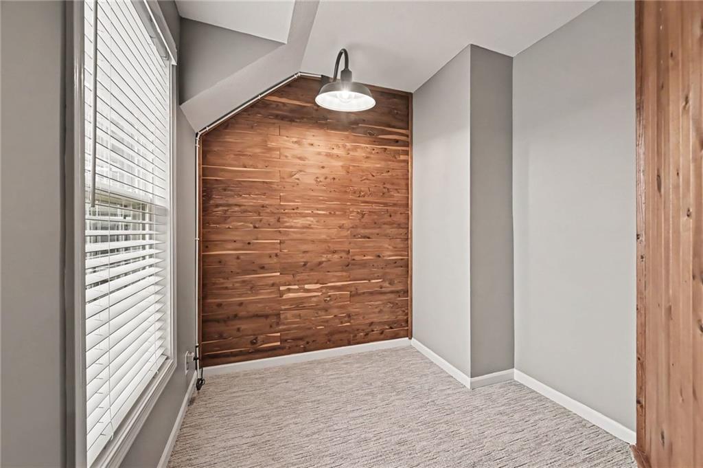 Vacant carpeted room with a rustic wood accent wall, tall window, and pendant light.