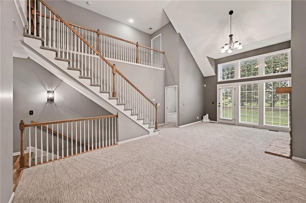 Vacant living room with a vaulted ceiling, sweeping staircase, and tall windows beside glass patio doors.