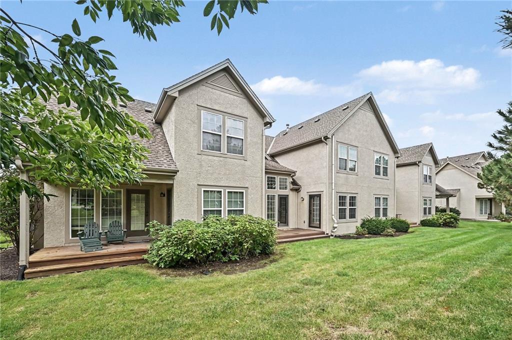 Rear of a beige multi-story home with a wooden deck, patio chairs, shrubs, and lawn.