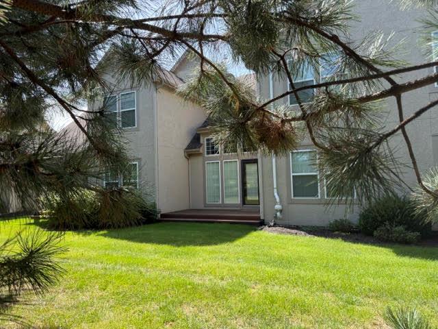 Rear of a two-story beige house with a small patio and pine branches framing the lawn.