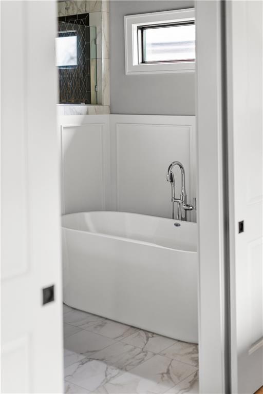 Bathroom with a freestanding white tub, chrome faucet, marble-patterned floor, and high window.