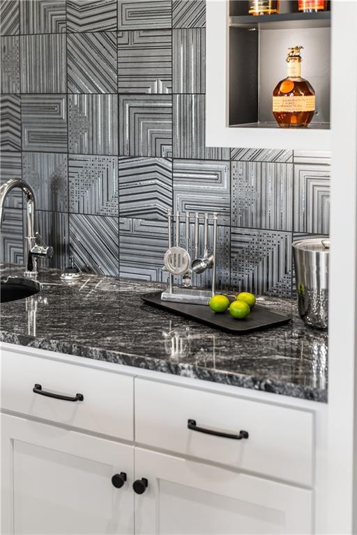 Wet bar with a geometric tile backsplash, dark stone counter, sink, and displayed spirits.