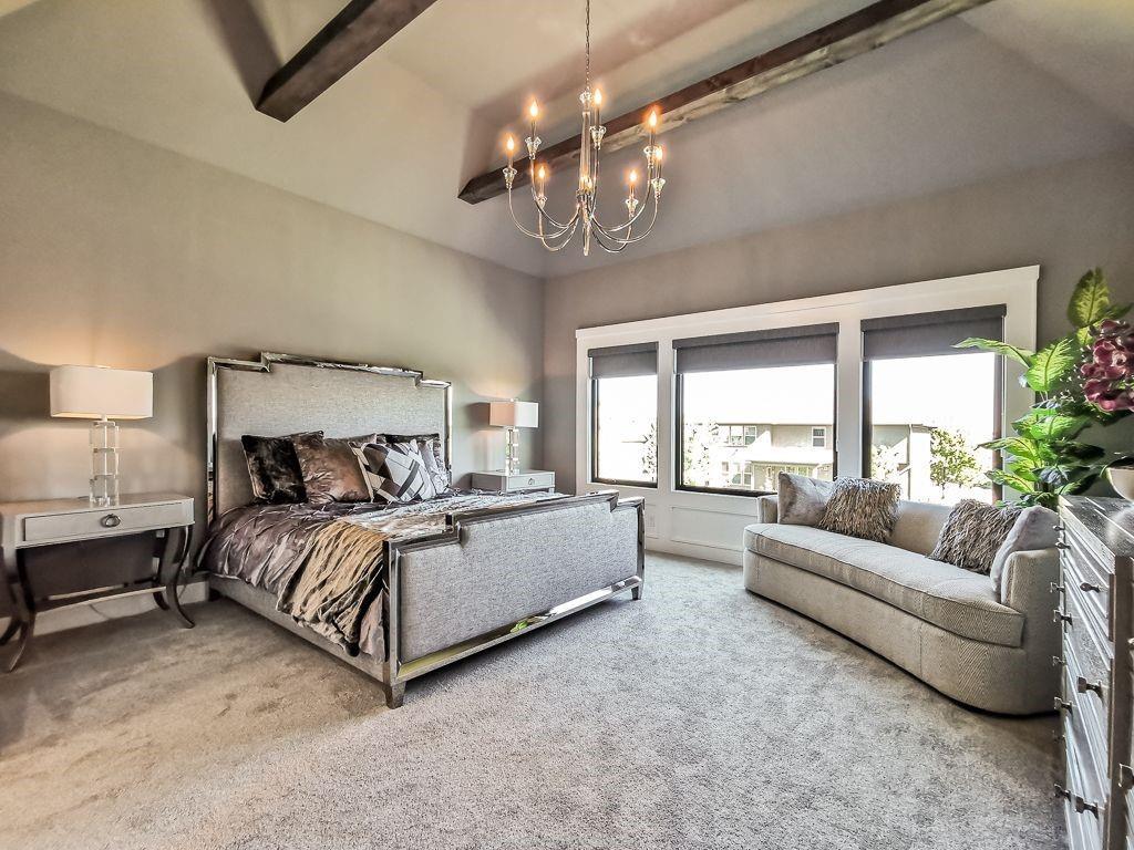 Staged bedroom with an upholstered bed, exposed ceiling beams, chandelier, and sofa by wide windows.