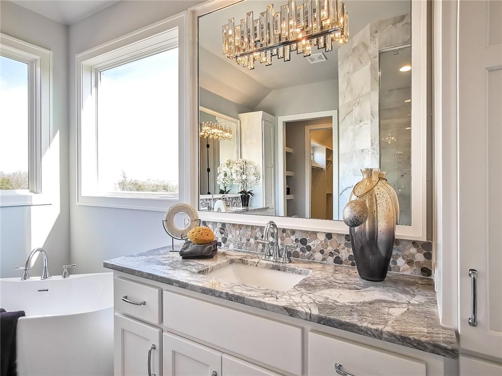 Bathroom vanity with marble countertop, mosaic backsplash, large mirror, and freestanding tub beside a window.