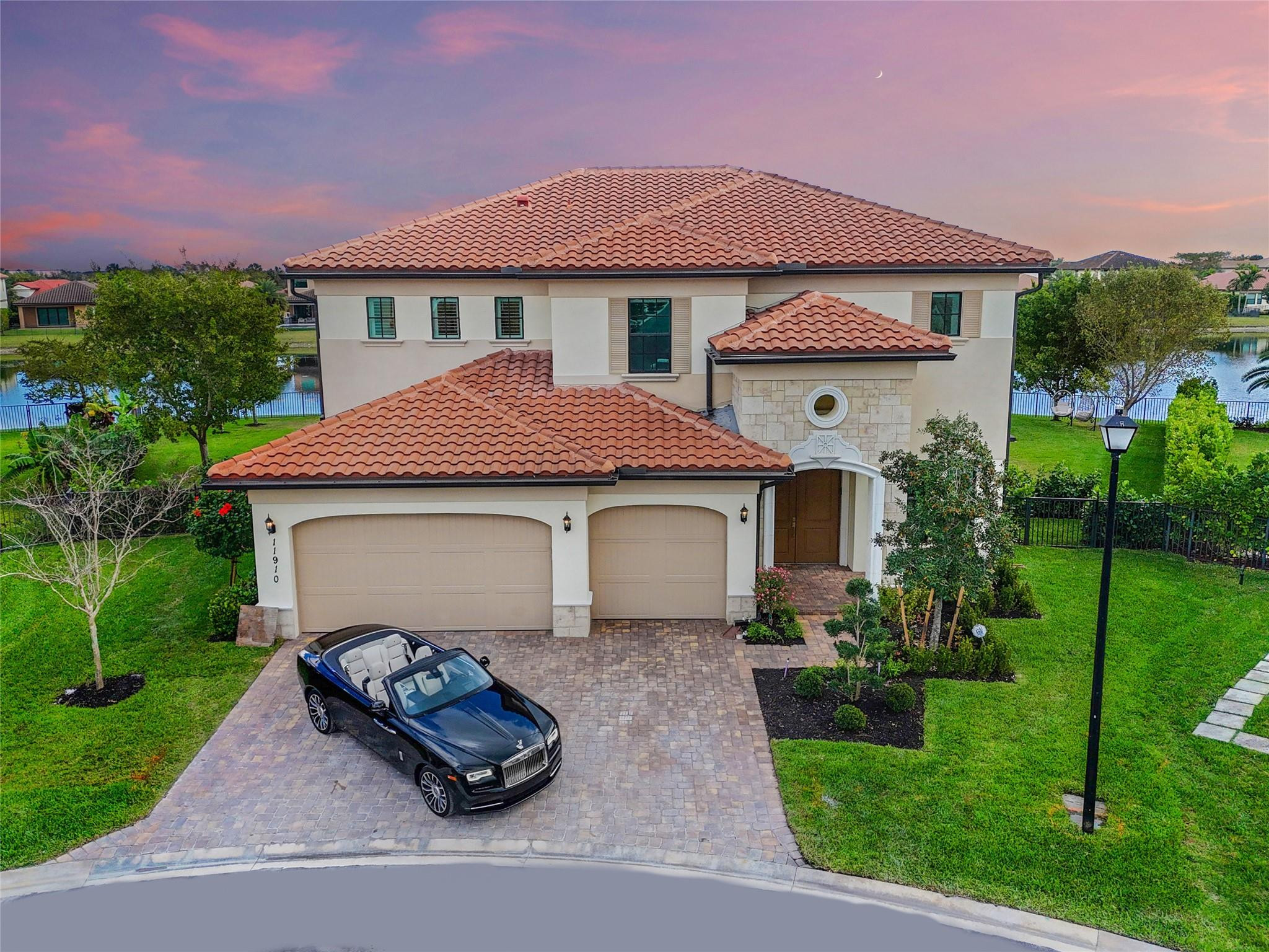 Aerial view of a two-story lakeside home with terracotta roofs and a black convertible in the driveway.