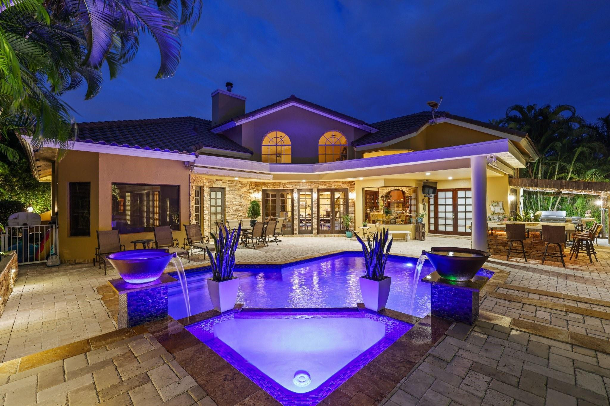 Two-story home and tropical patio at night, with a purple-lit pool and decorative water bowls.