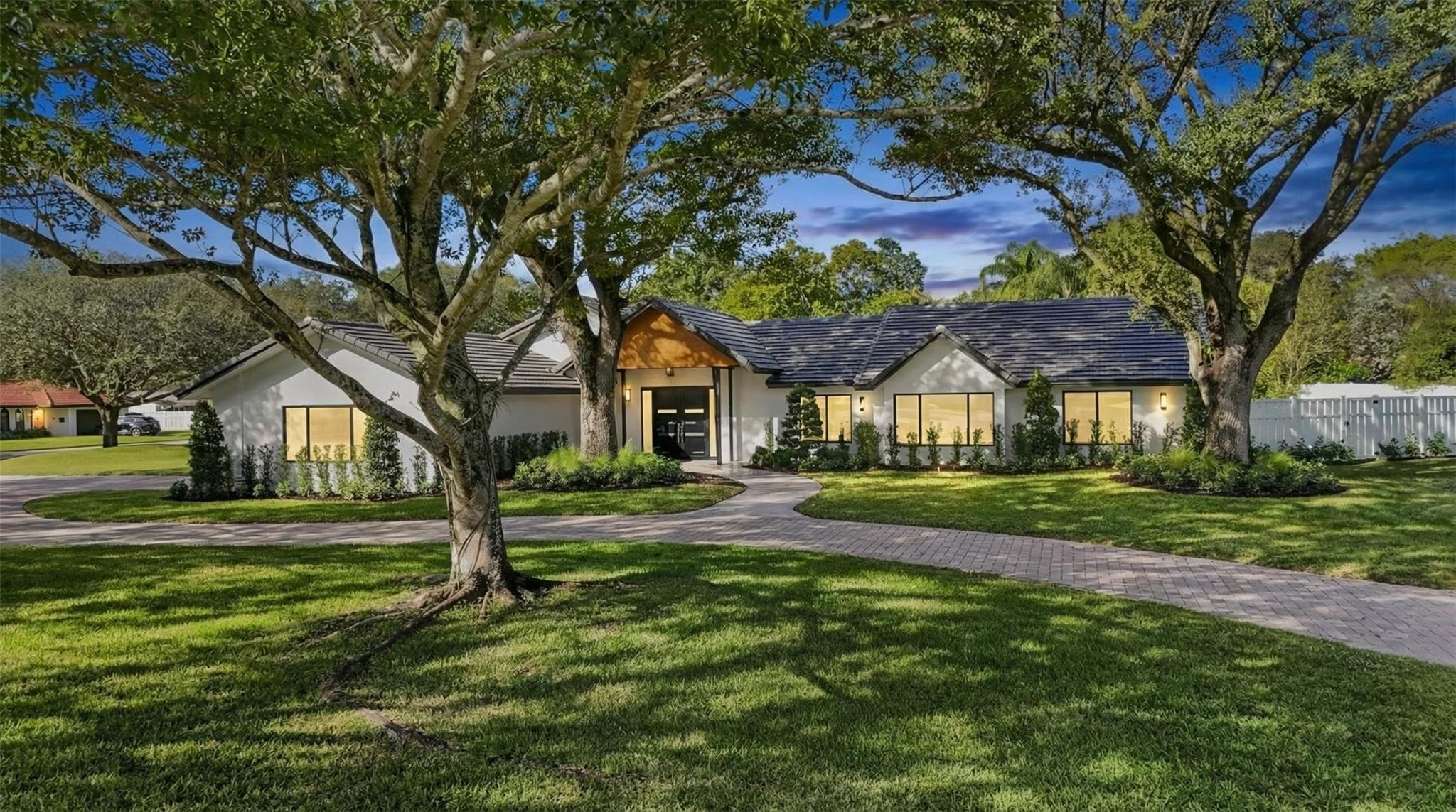 Single-story white home with a dark tiled roof, framed by mature trees and a curved brick walkway.