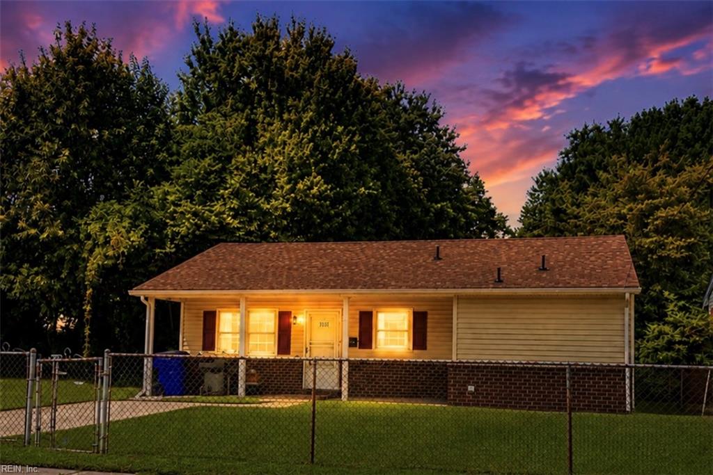 Single-story ranch house with glowing windows behind a chain-link fence at dusk.