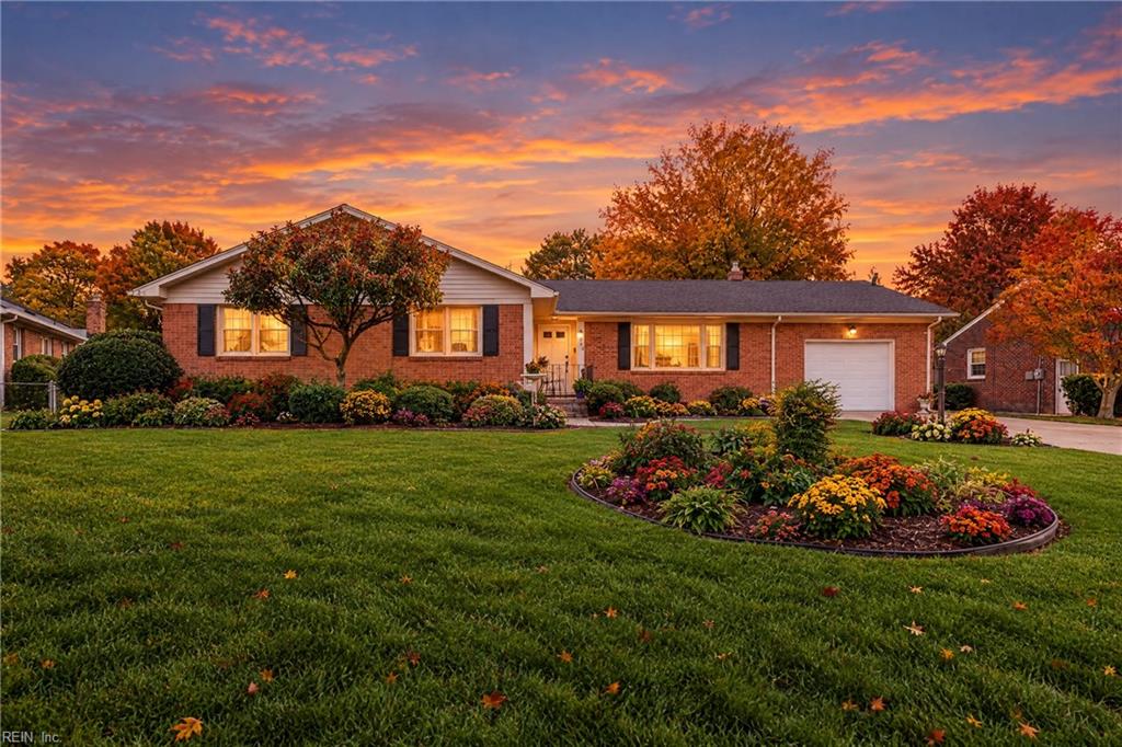 Brick ranch home with an attached garage, flower beds, and a broad lawn beneath a colorful sunset.