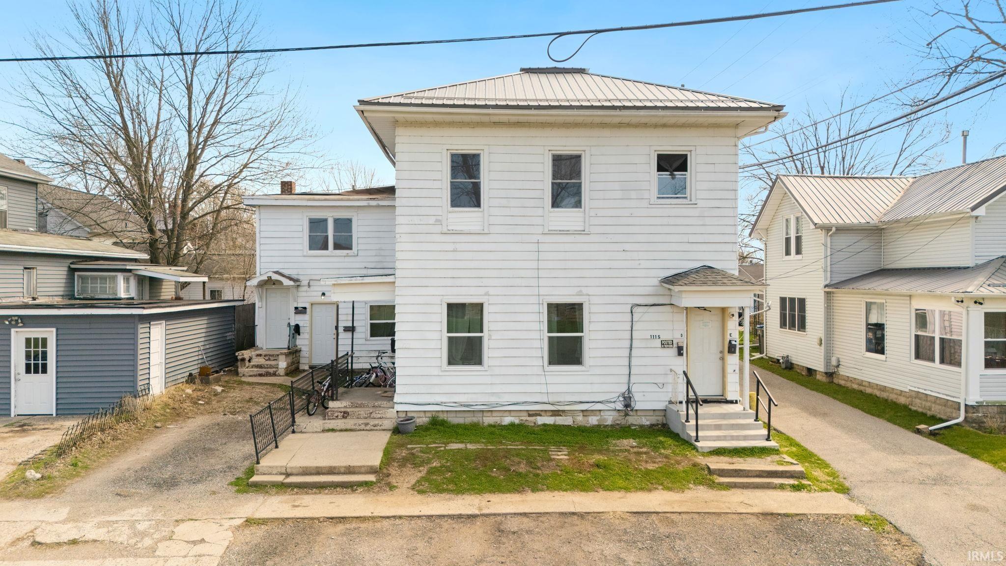 White two-story house with a metal roof, front steps, and neighboring homes close on both sides.
