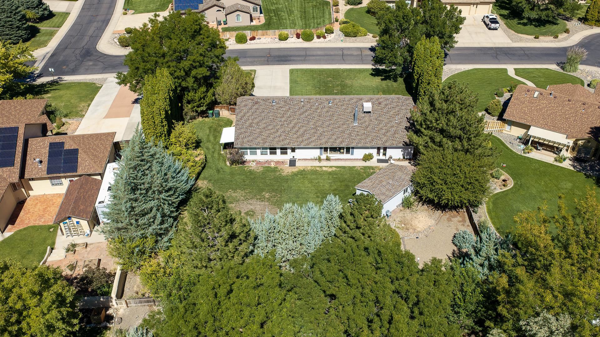 Aerial view of a single-story home with a broad lawn, mature trees, and a small backyard outbuilding.