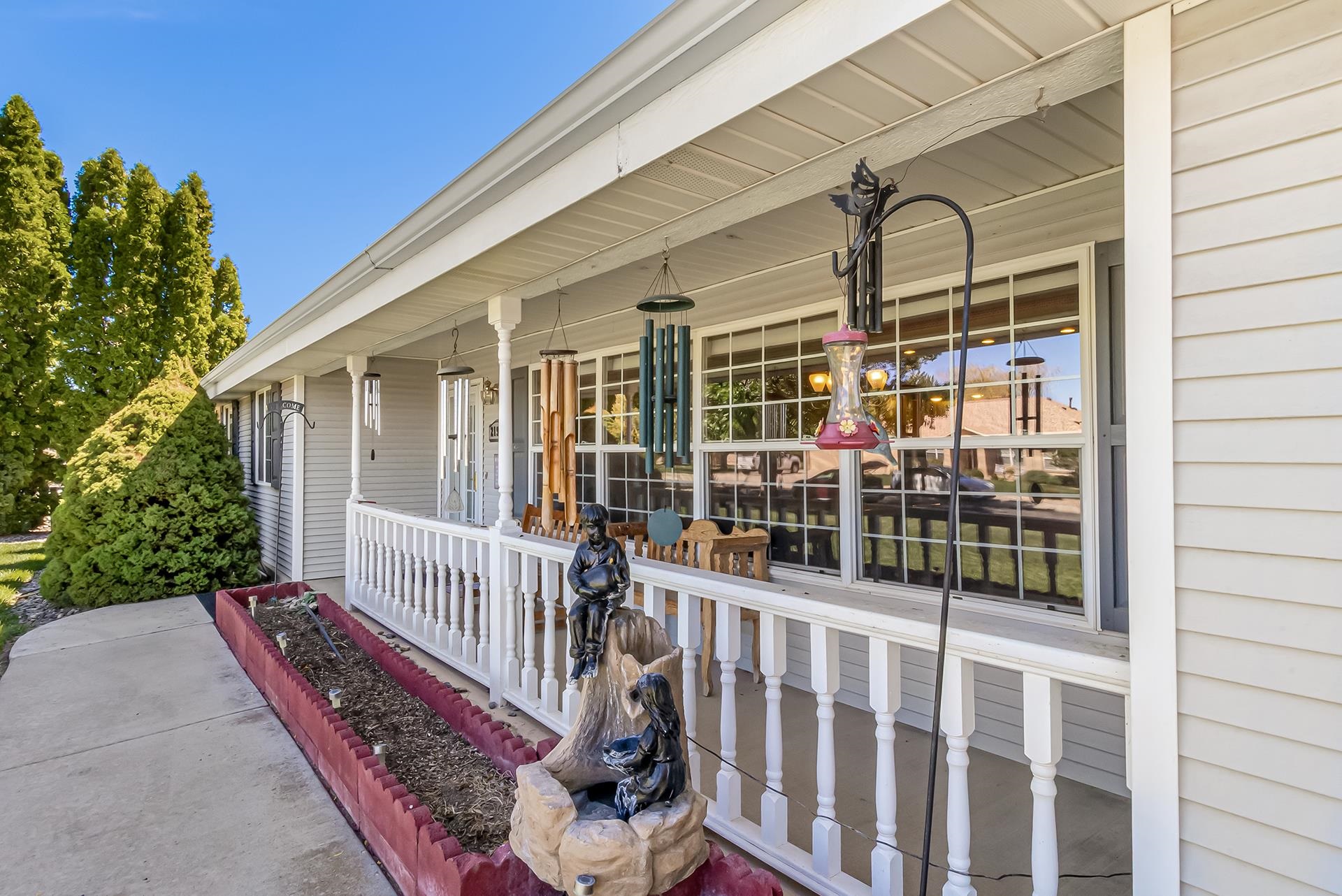 White home with a covered front porch, large windows, hanging wind chimes, and garden sculptures.