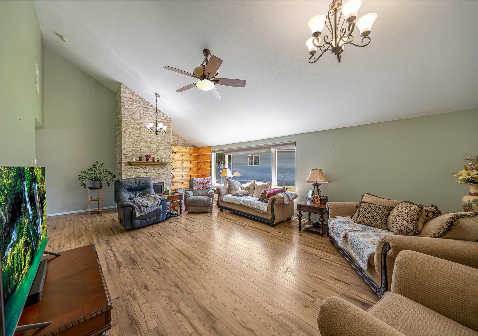 Living room with vaulted ceiling, stone fireplace, large windows, and wood-look flooring.