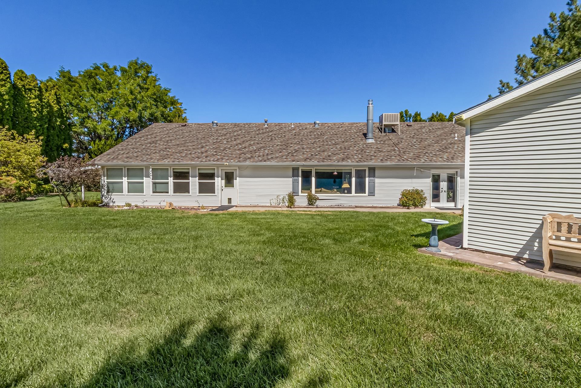 Rear of a single-story home with a broad lawn, mature trees, and a small patio area.