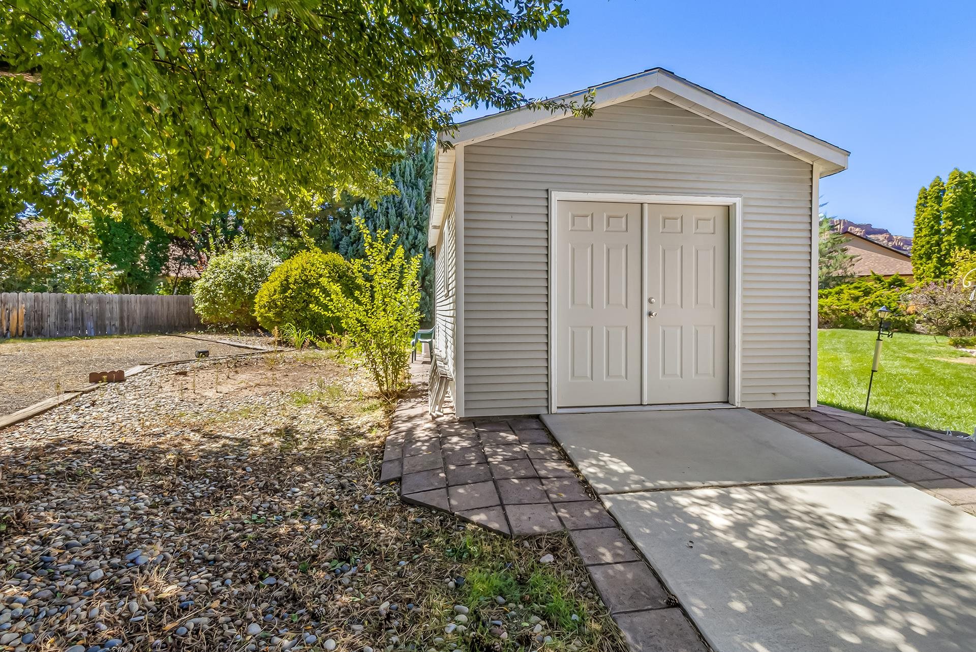 Detached light-colored outbuilding with double doors beside a paved driveway and landscaped yard.