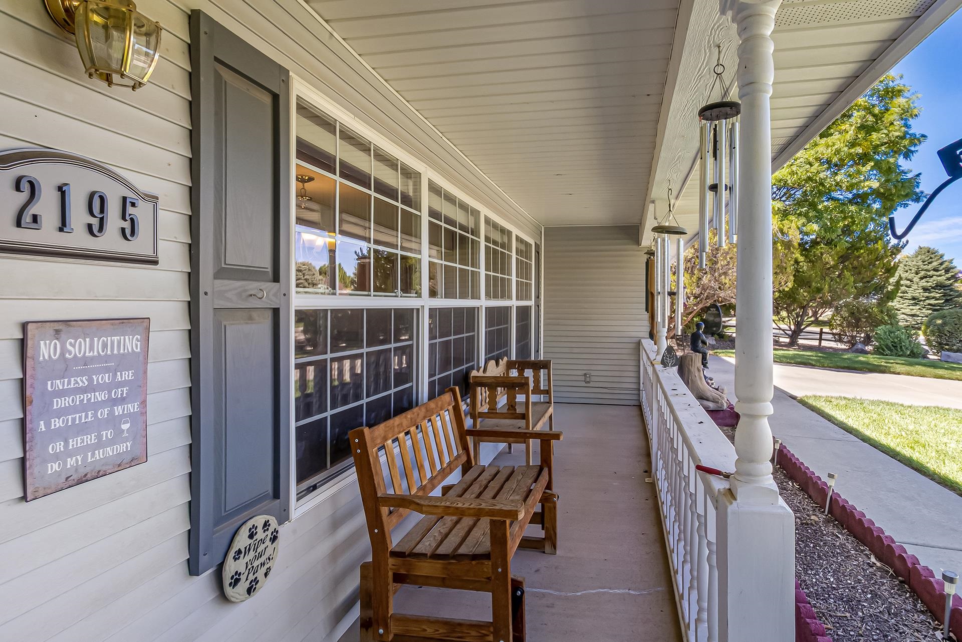Covered front porch with wooden benches, screened windows, and wind chimes overlooking a sunny landscaped yard.