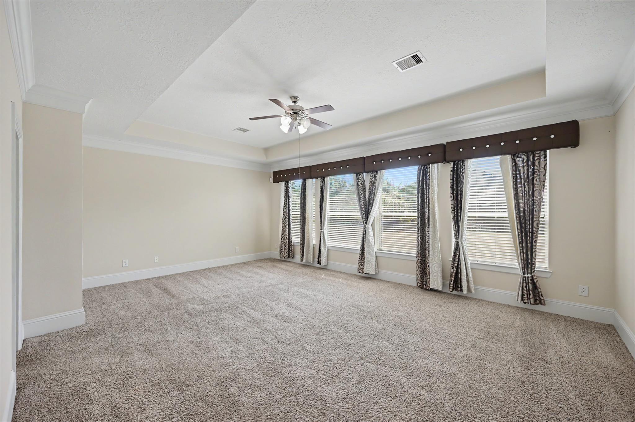 Empty carpeted room with a tray ceiling, ceiling fan, and curtained windows.