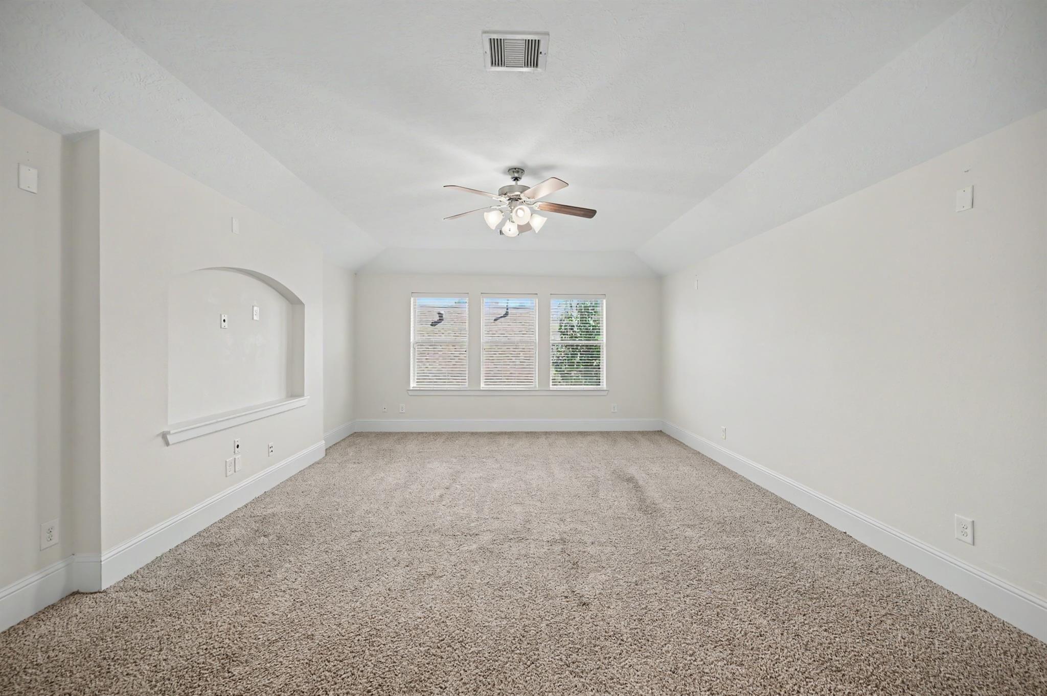 Empty carpeted room with three shuttered windows, an arched wall niche, and a ceiling fan.