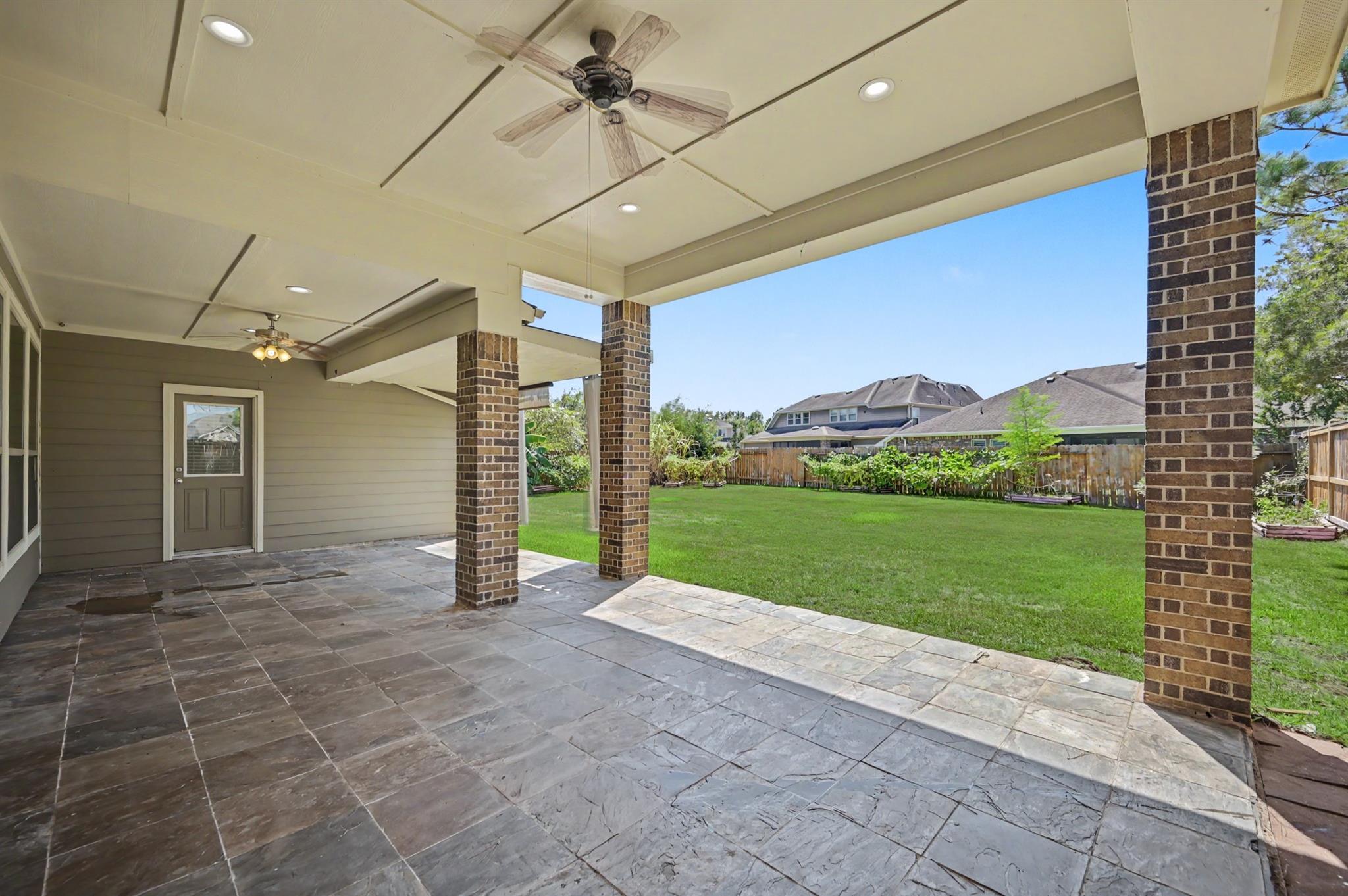 Covered patio with stone tile flooring and brick columns overlooking a fenced backyard.