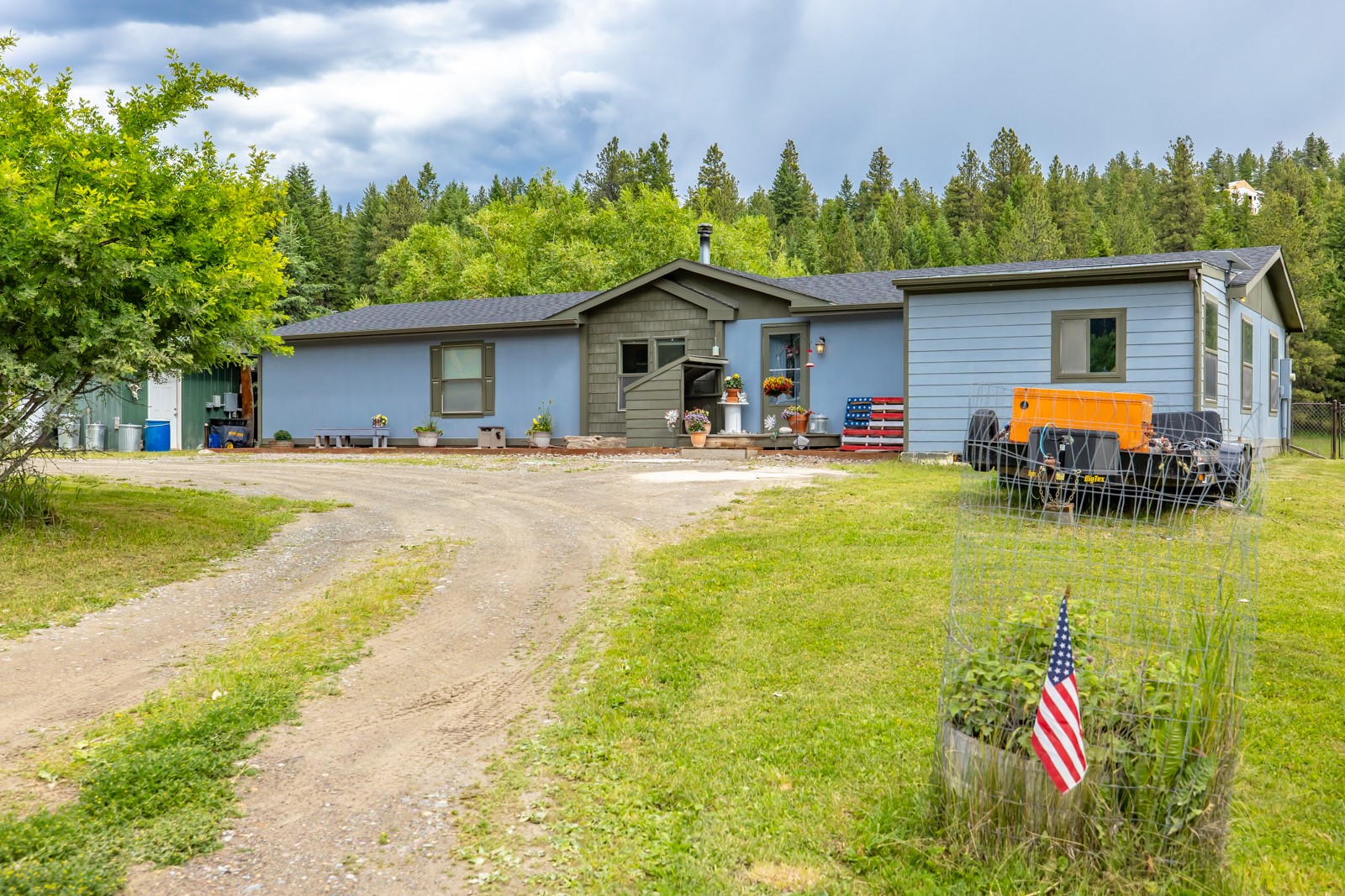 Blue single-story home with a central covered entry, gravel driveway, and wooded hillside behind it.