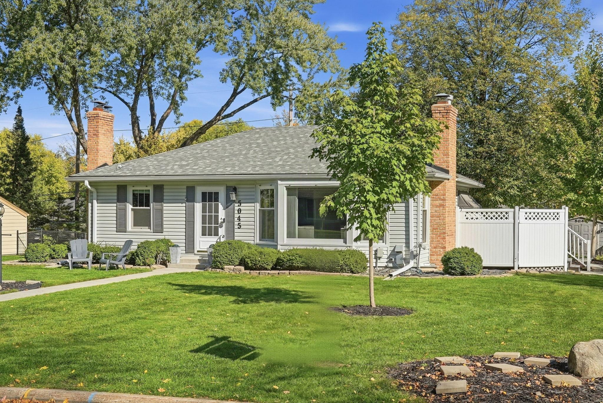 Gray single-story house with twin brick chimneys, a broad lawn, outdoor chairs, and a white fence.