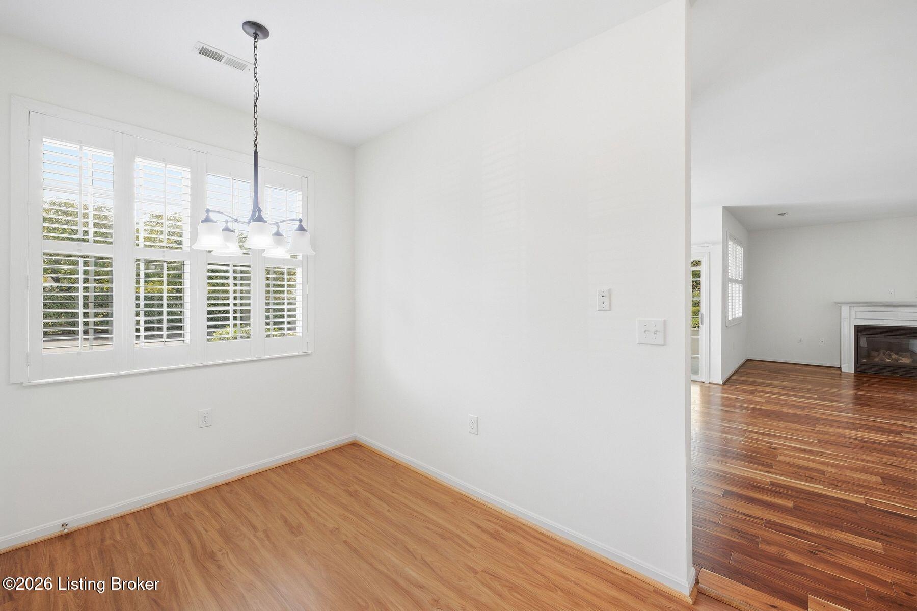 Vacant dining area with plantation shutters, a hanging chandelier, and an adjoining living room with fireplace.