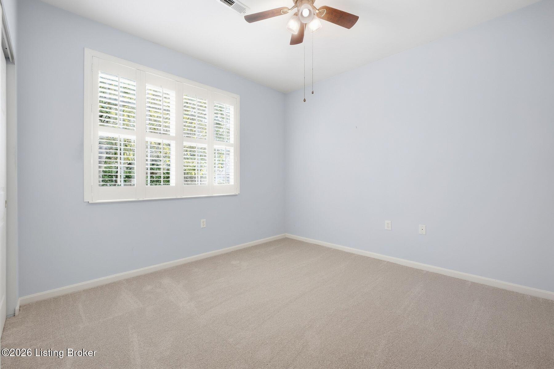 Empty carpeted room with pale blue walls, plantation shutters, and a ceiling fan with light.