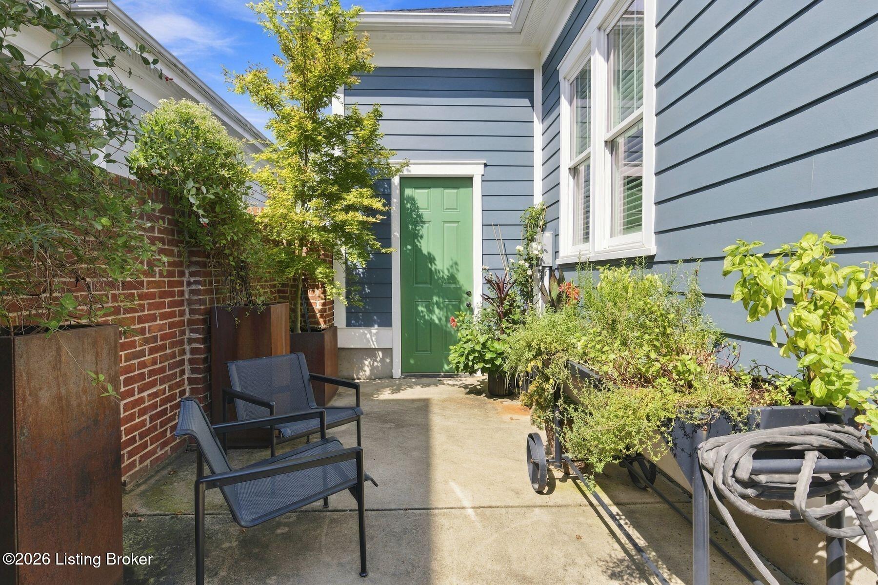 Patio beside a blue-sided home with a green entry door, black chairs, and potted plants.