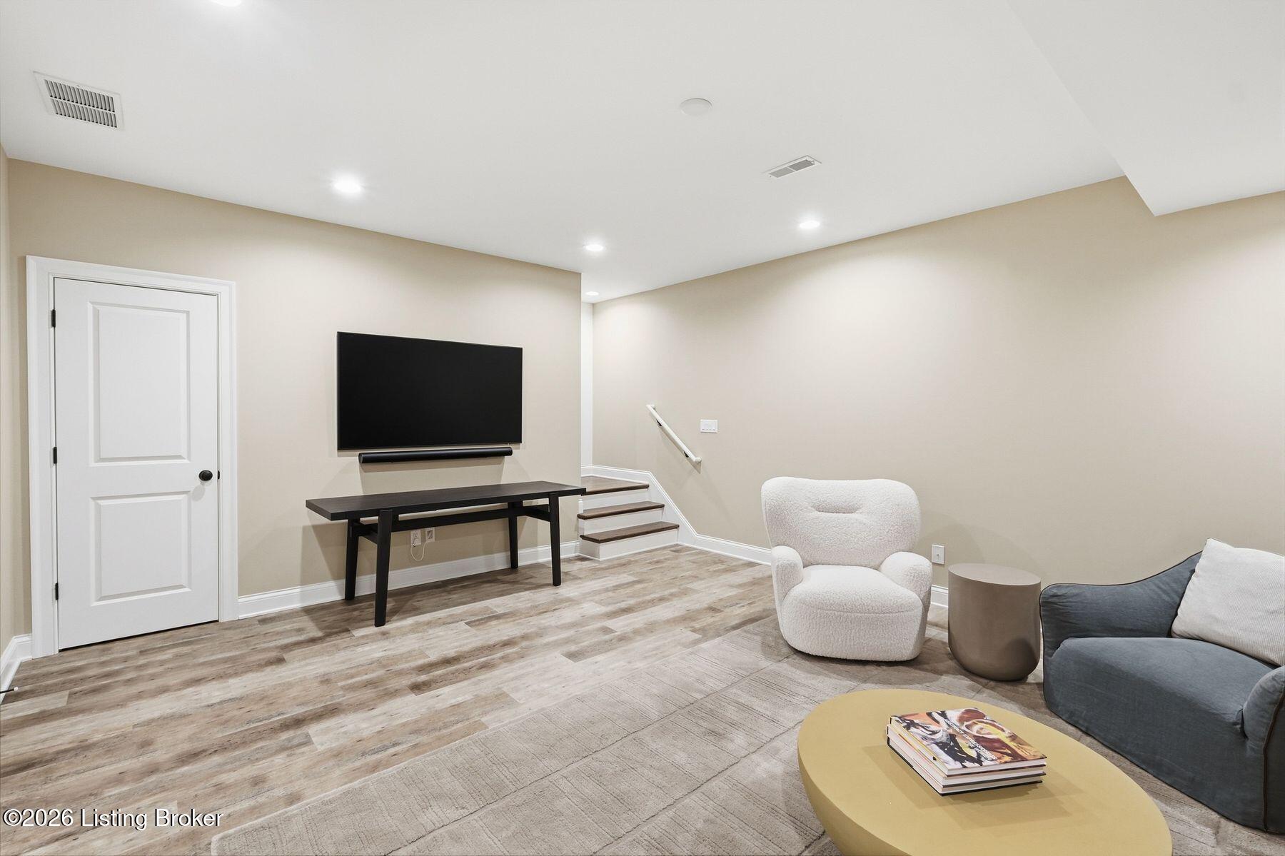 Basement living area with a wall-mounted TV, white chair, gray sofa, and stairs.