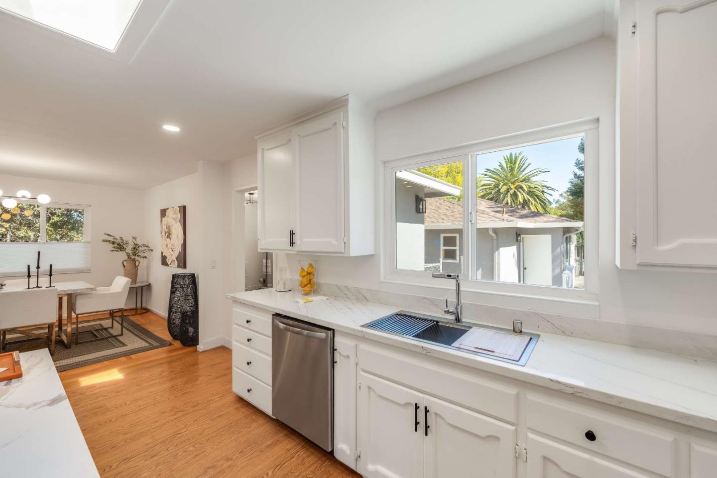 Bright white kitchen with marble counters, stainless dishwasher, and a window overlooking a palm tree.
