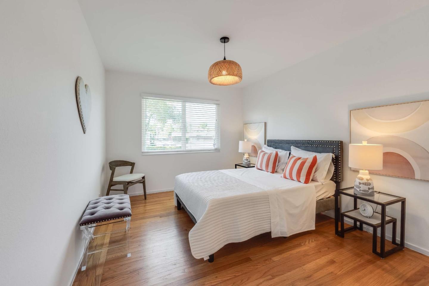 Staged bedroom with a white bed, coral-striped pillows, hardwood floors, and a window with blinds.