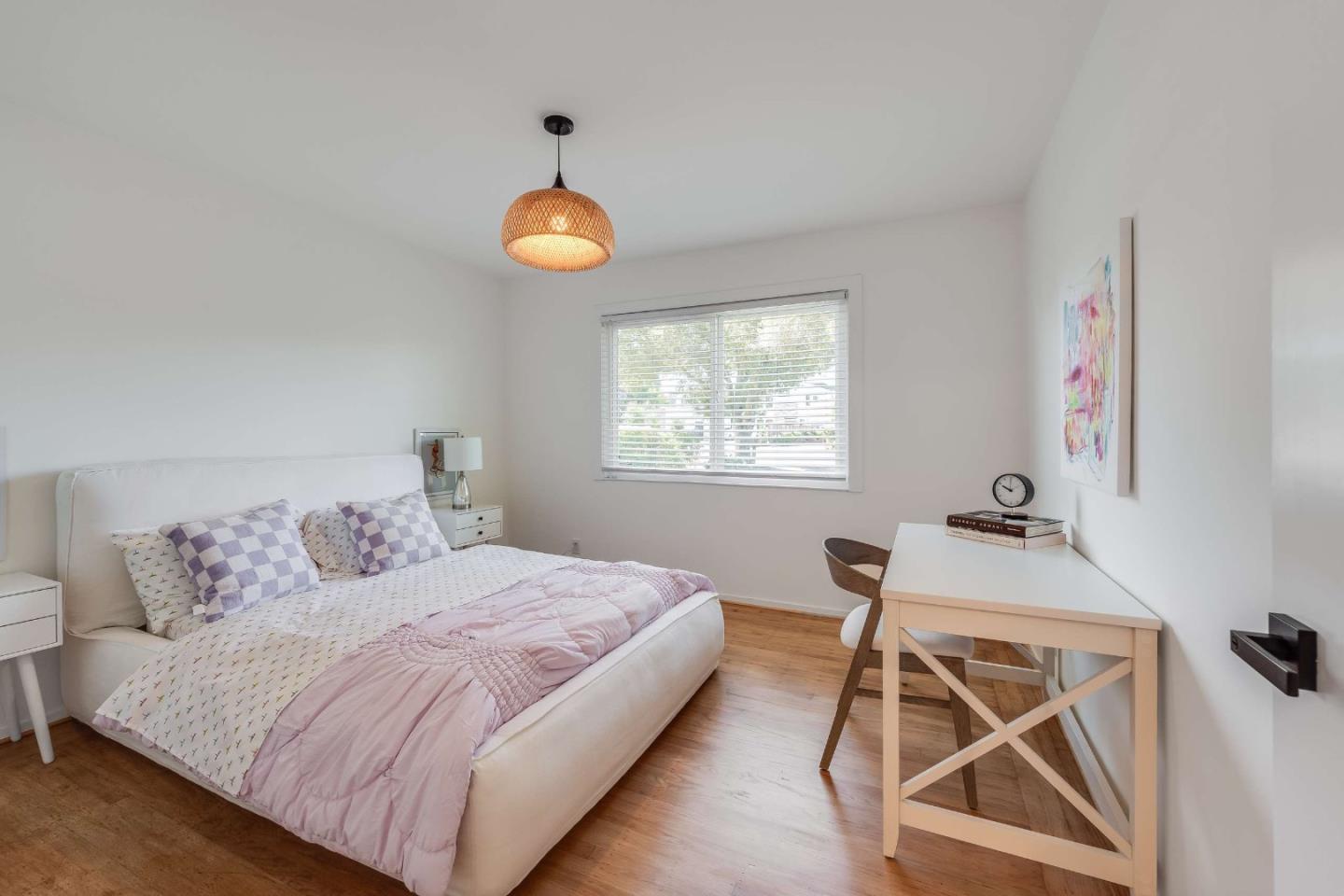 Bright bedroom with a white bed, pink and lavender bedding, hardwood floors, and a desk by the window.