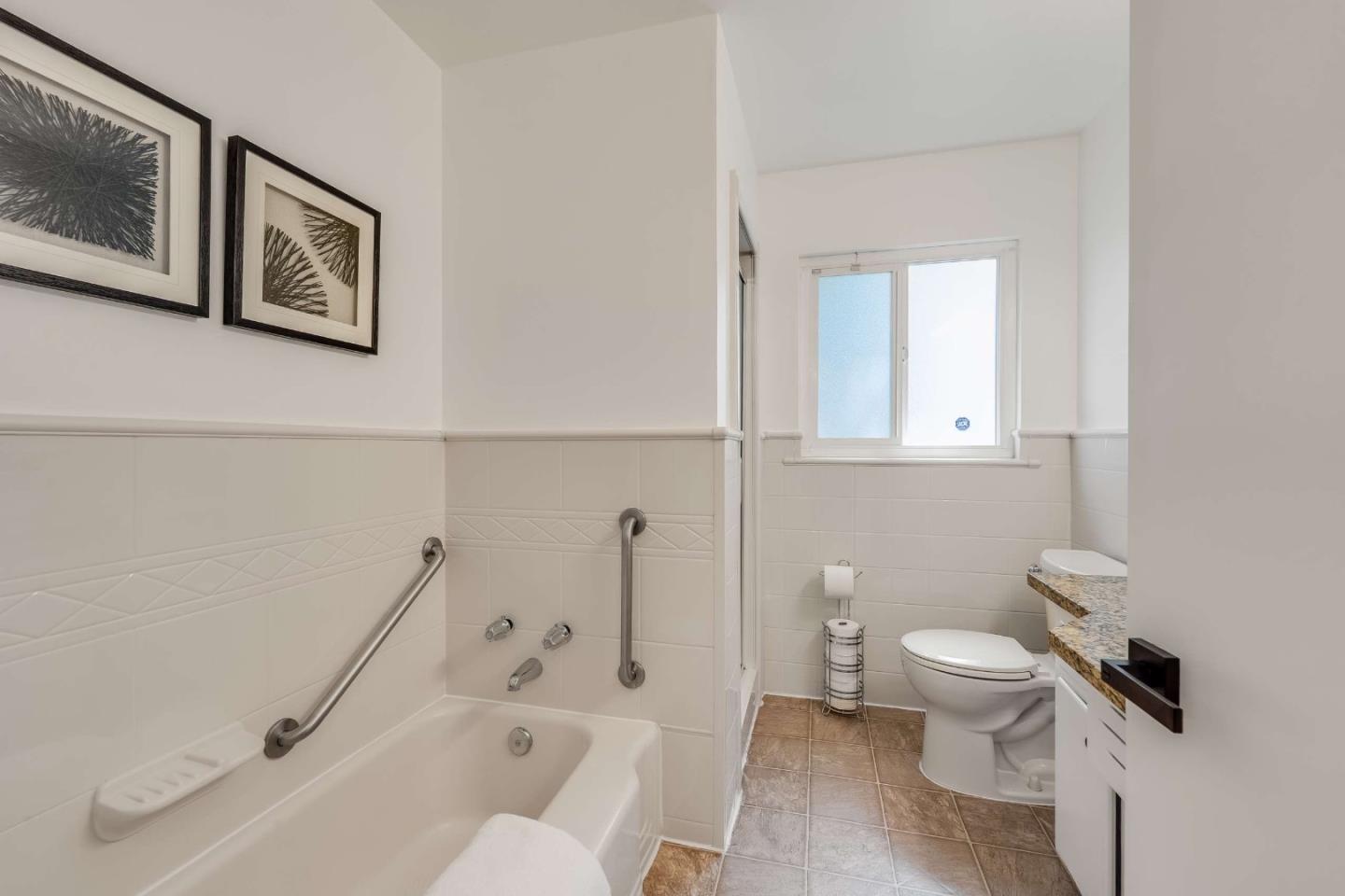 Bathroom with a grab-bar-equipped tub, toilet, granite vanity, and frosted window.