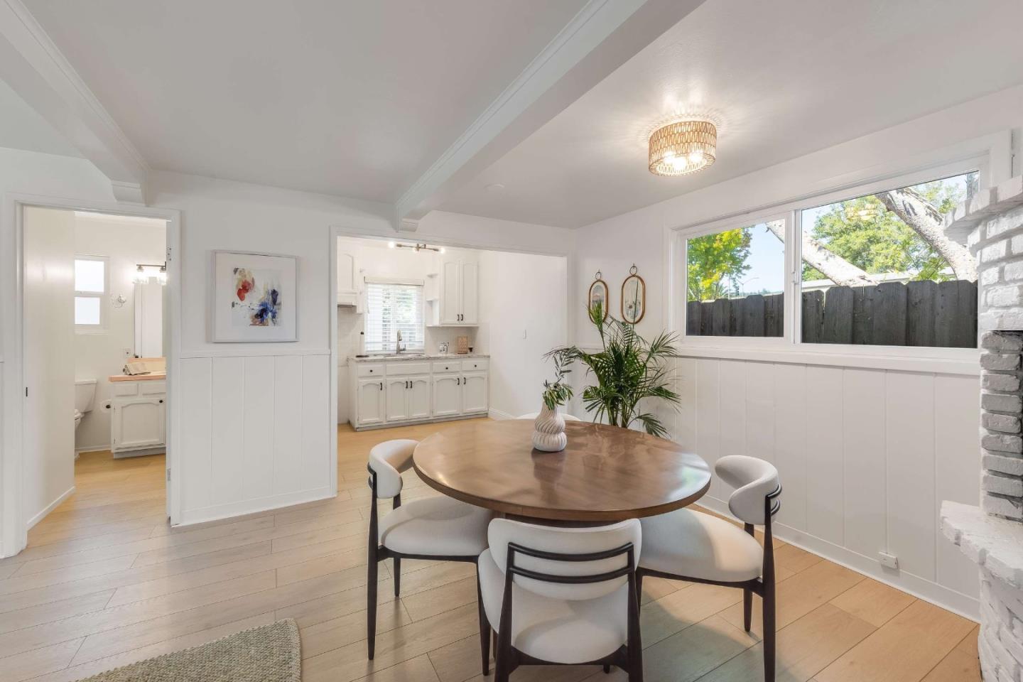 Dining room with a round wooden table, white kitchen, large yard-facing window, and brick fireplace.
