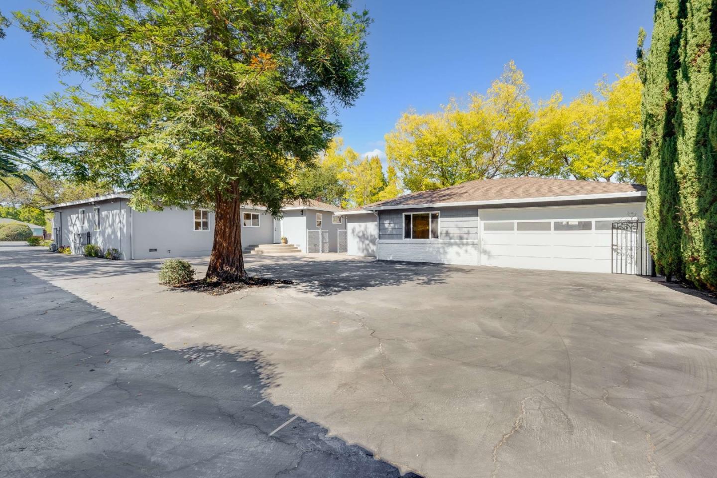 Single-story gray house with a two-car garage, broad driveway, mature trees, and yellow autumn foliage.