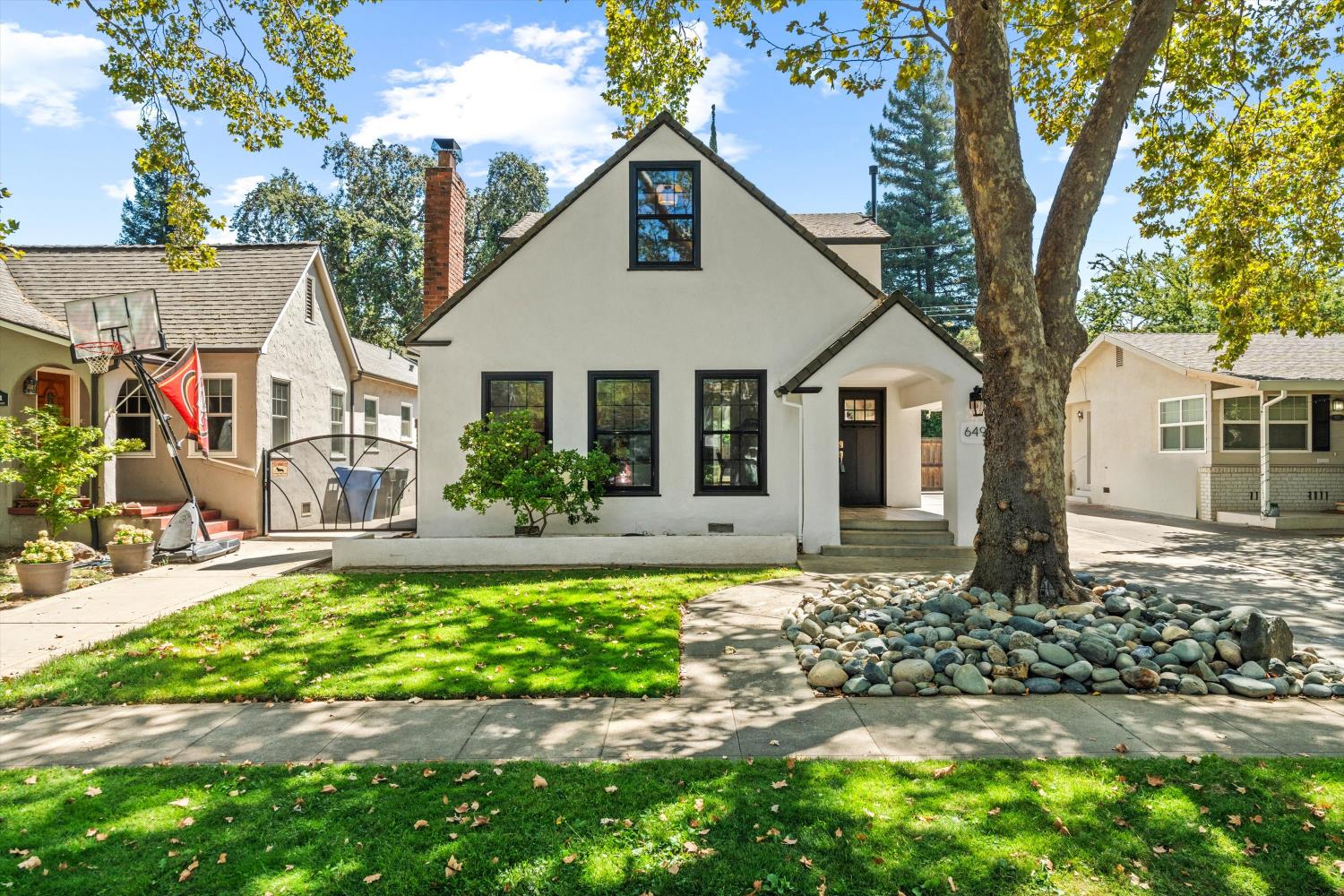 White stucco house with black-trimmed windows, a mature front-yard tree, lawn and rock landscaping.