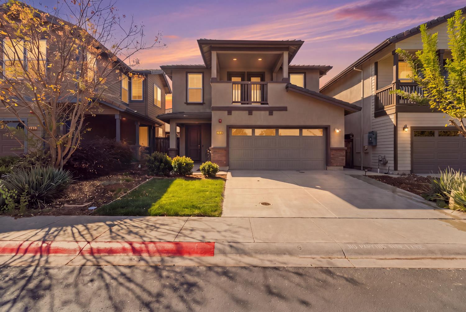Two-story home with a balcony and double garage, photographed at sunset among neighboring houses.
