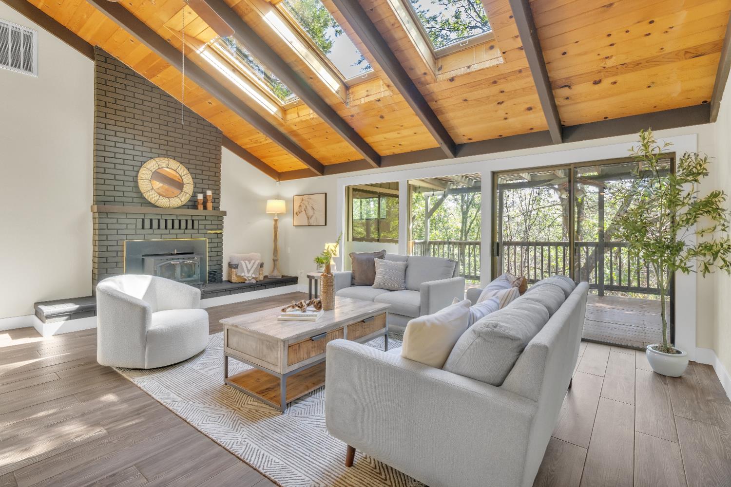 Living room with a vaulted wood ceiling, skylights, brick fireplace, and glass doors to a tree-lined deck.