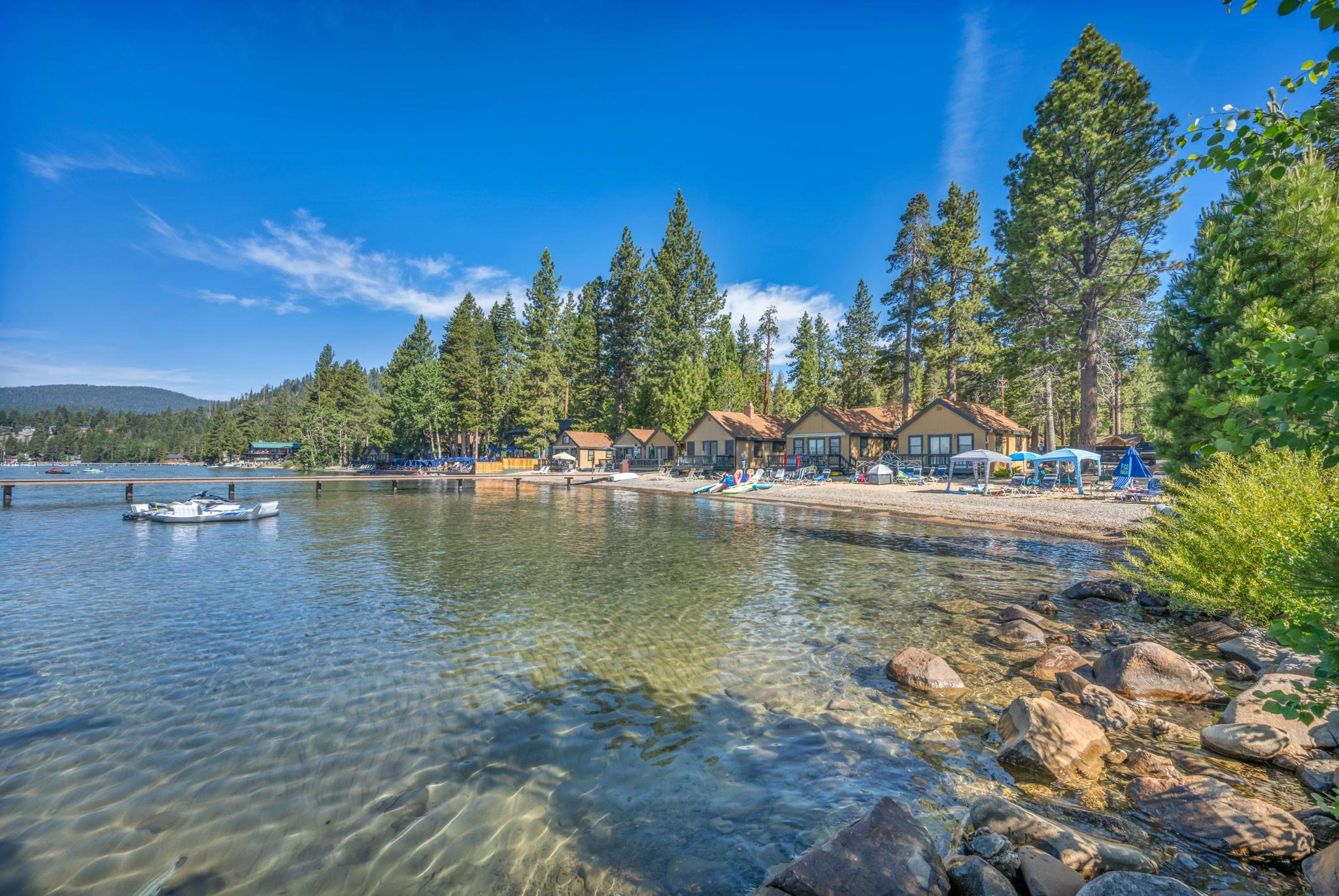 Lakeside cabins line a clear rocky shore beside a wooden dock and pine trees.