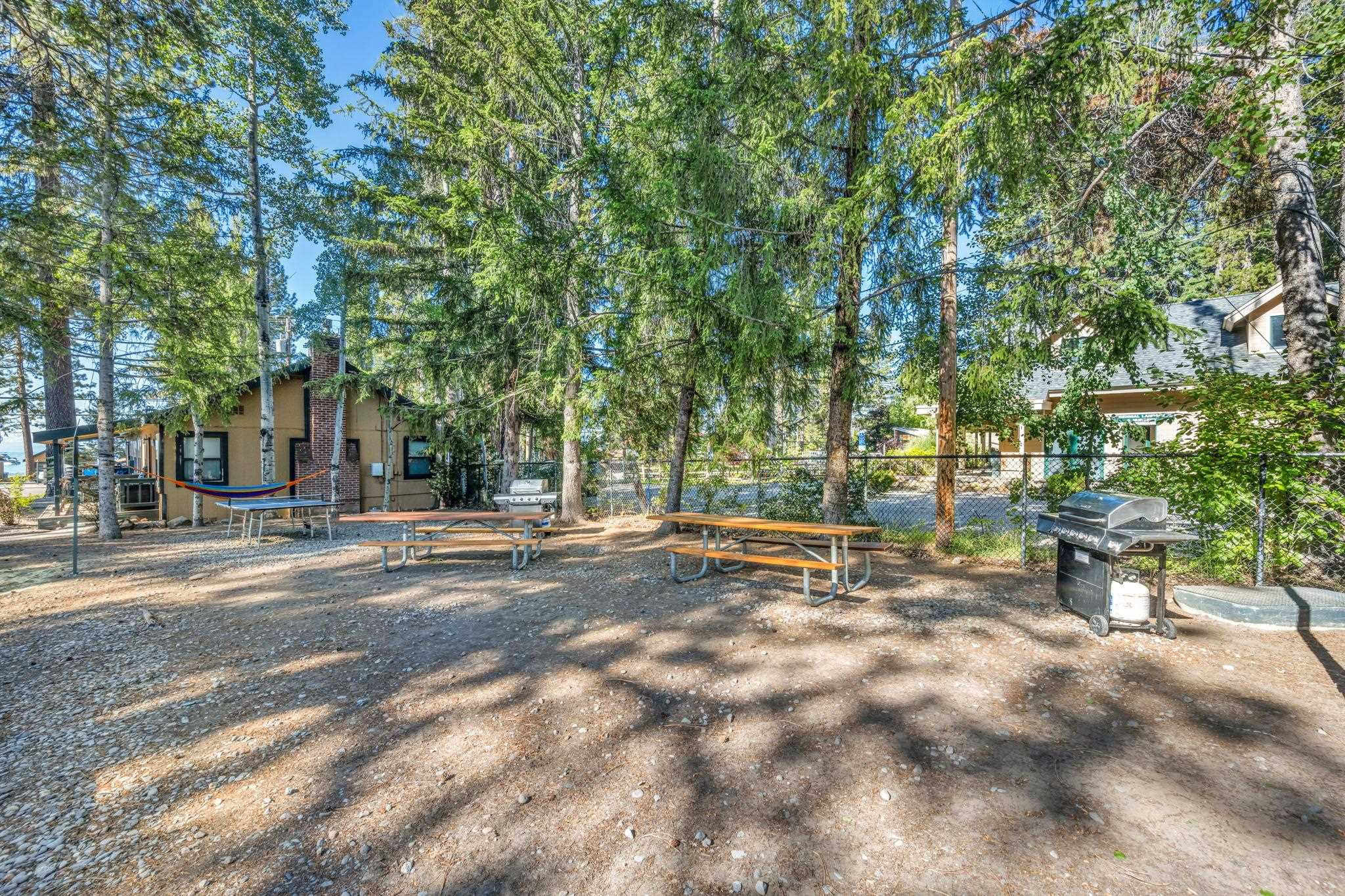 Wooded outdoor picnic area with picnic tables, a hammock, and a barbecue grill beside residential buildings.