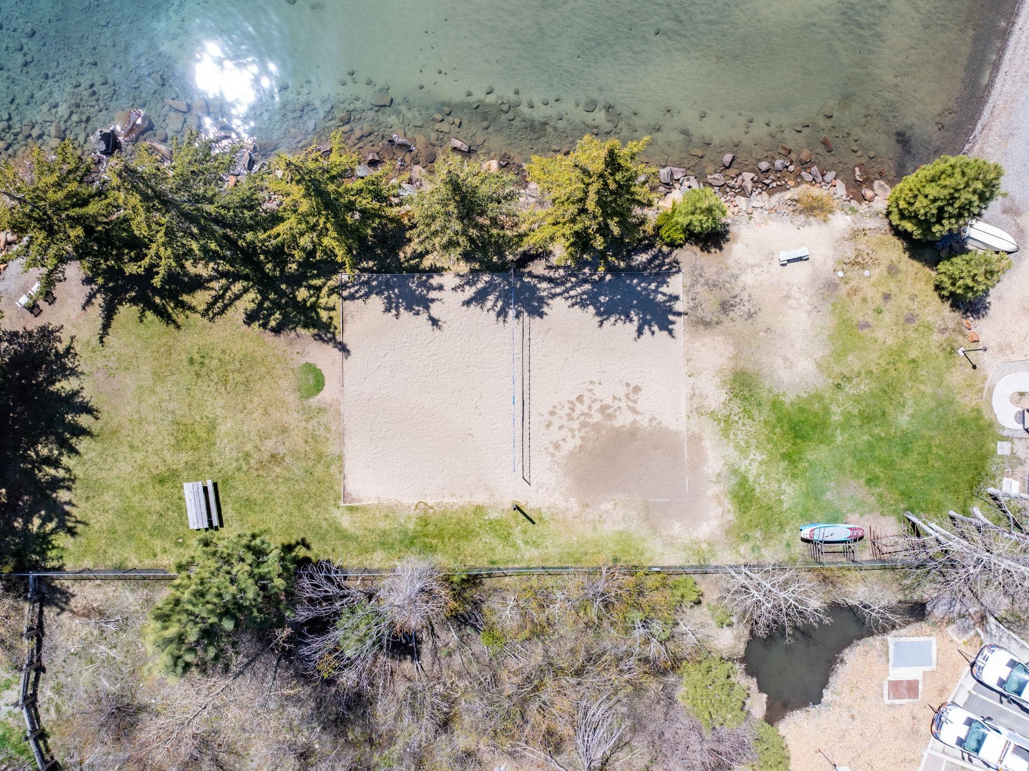 Aerial view of a sandy volleyball court beside a rocky lakeshore, with trees and grassy recreation areas.