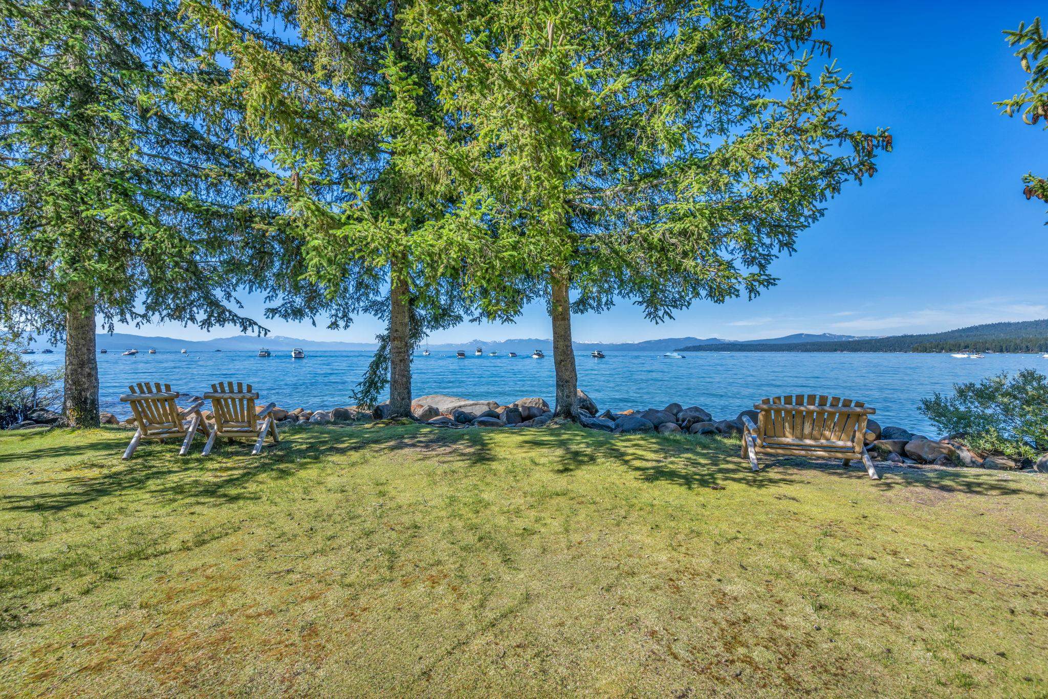 Lakeside lawn with Adirondack chairs beneath evergreen trees, overlooking boats and distant mountains.