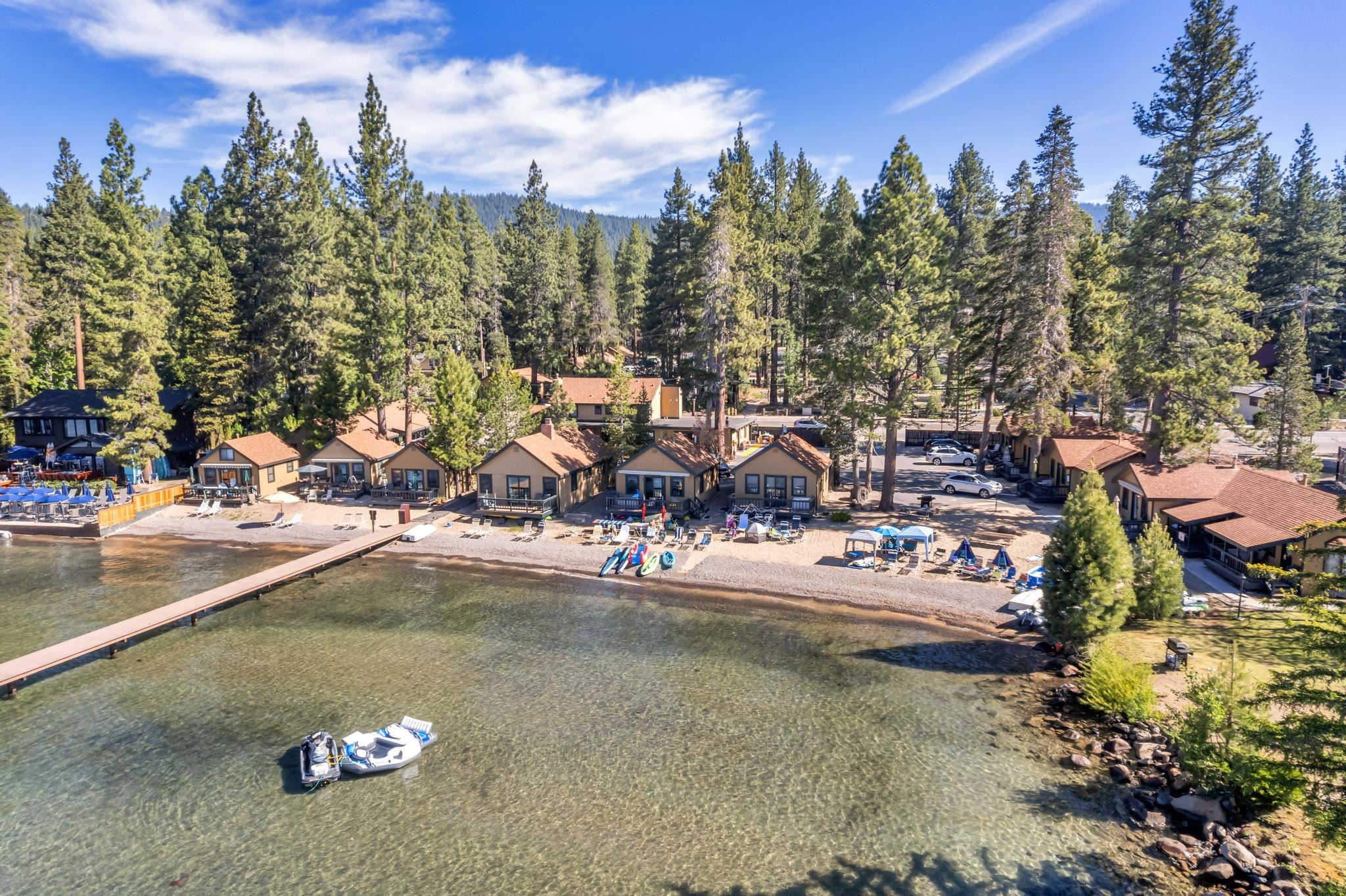 Lakeside cottages line a pebbled beach beside a long pier, kayaks, and a boat.