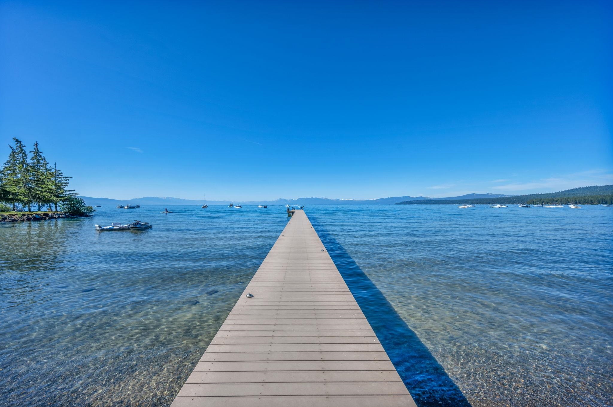 A long wooden dock extends into a clear lake, with boats and distant mountains.