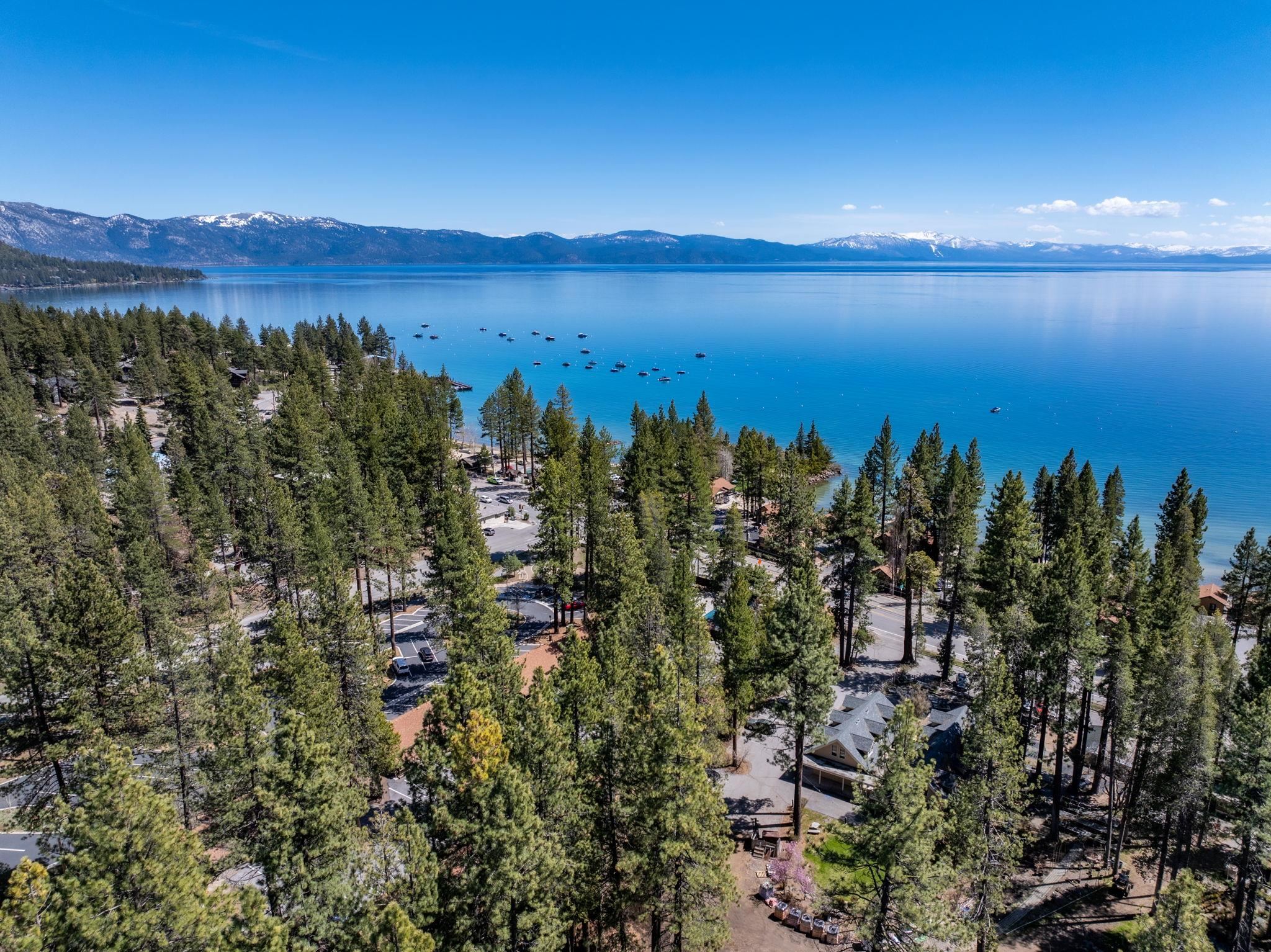 Aerial view of a pine-lined lakeshore, blue lake, boats, and distant snow-dusted mountains.