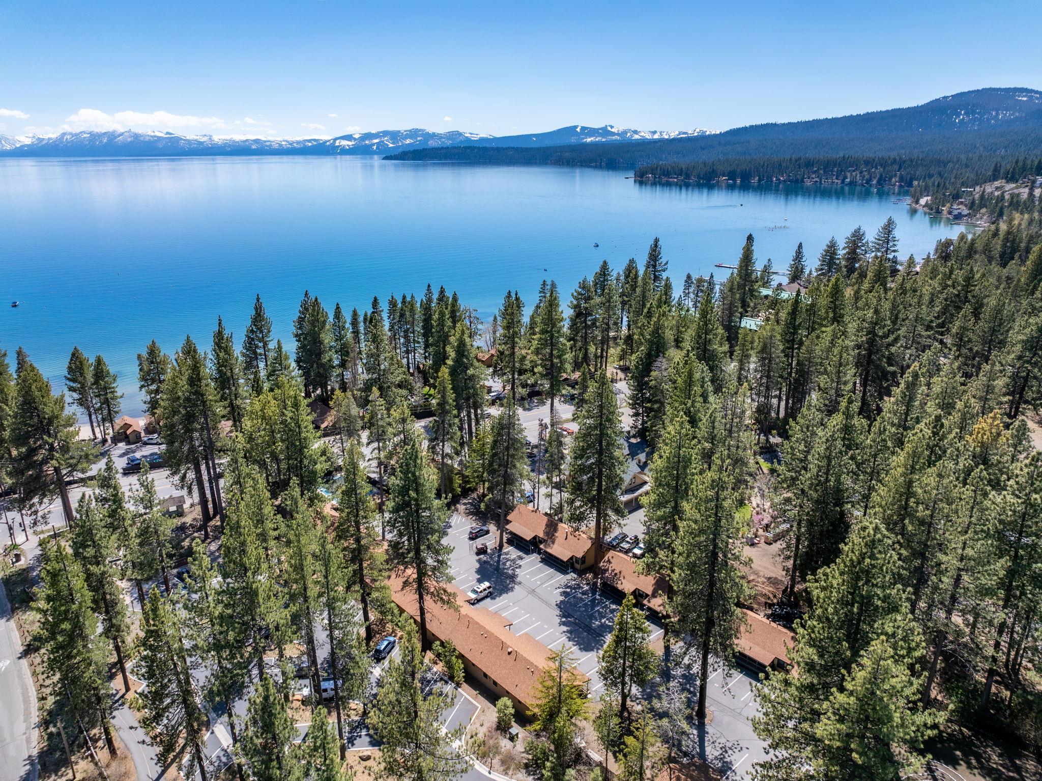 Aerial view of lakeside condominiums among tall pines, with blue water and snow-dusted mountains beyond.