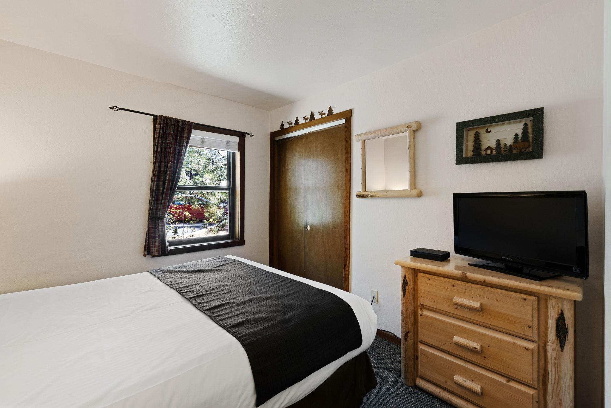 Bedroom with a white bed, dark runner, plaid-curtained window, and rustic wooden dresser with television.