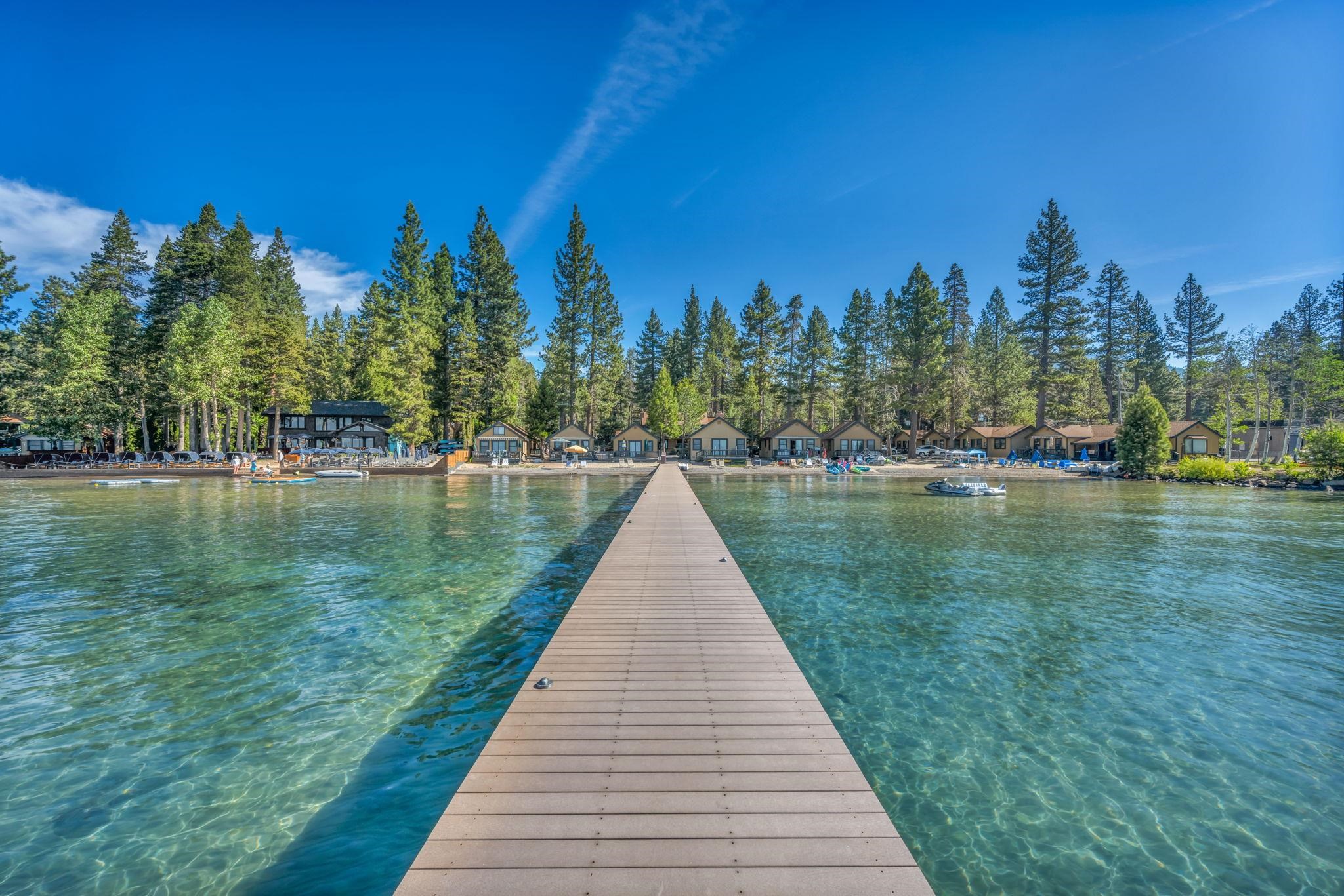 Wooden dock crosses clear lake water toward lakeside cabins and tall pine trees.