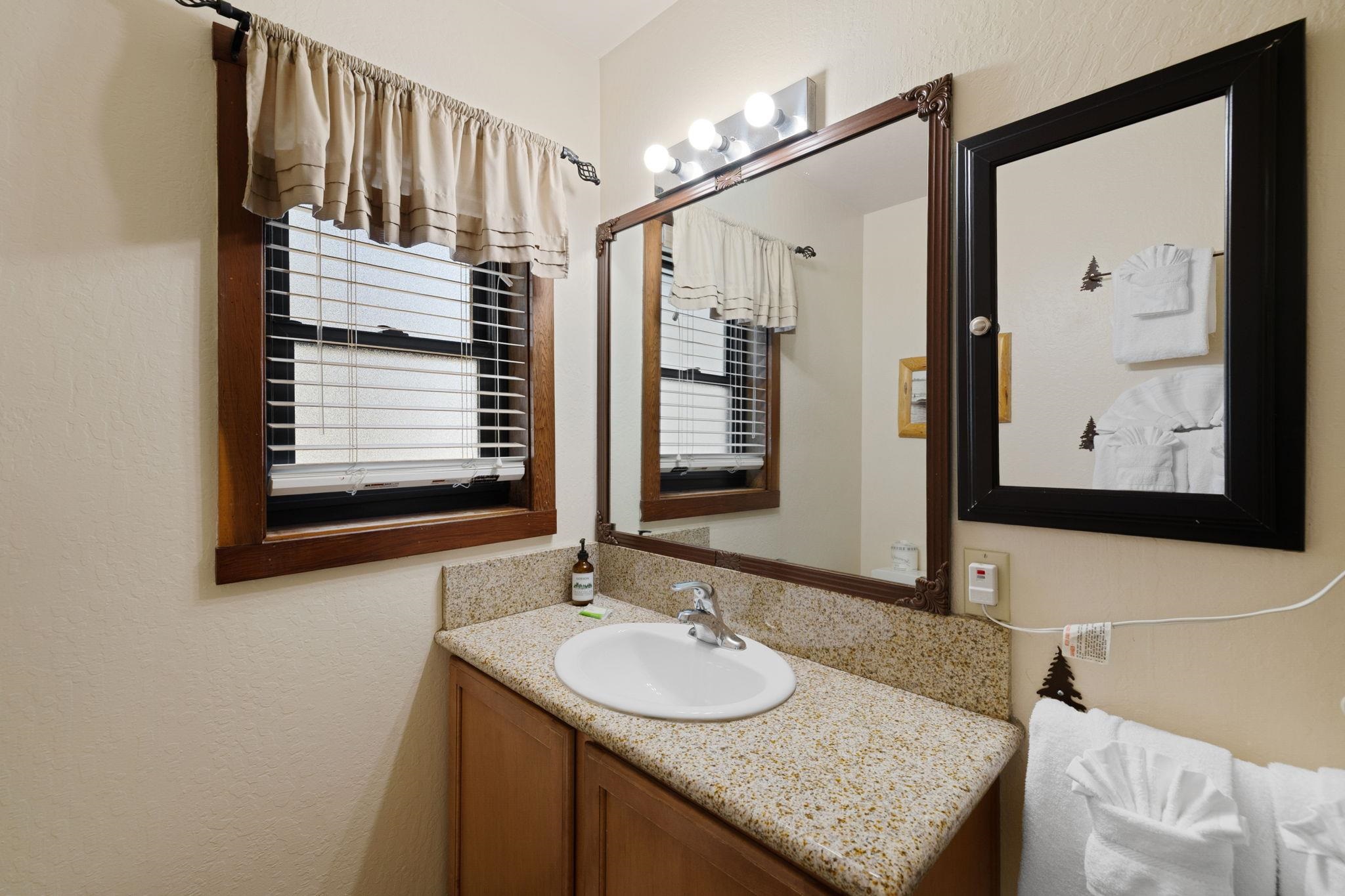 Bathroom vanity with speckled countertop, oval sink, large framed mirror, and window with blinds.