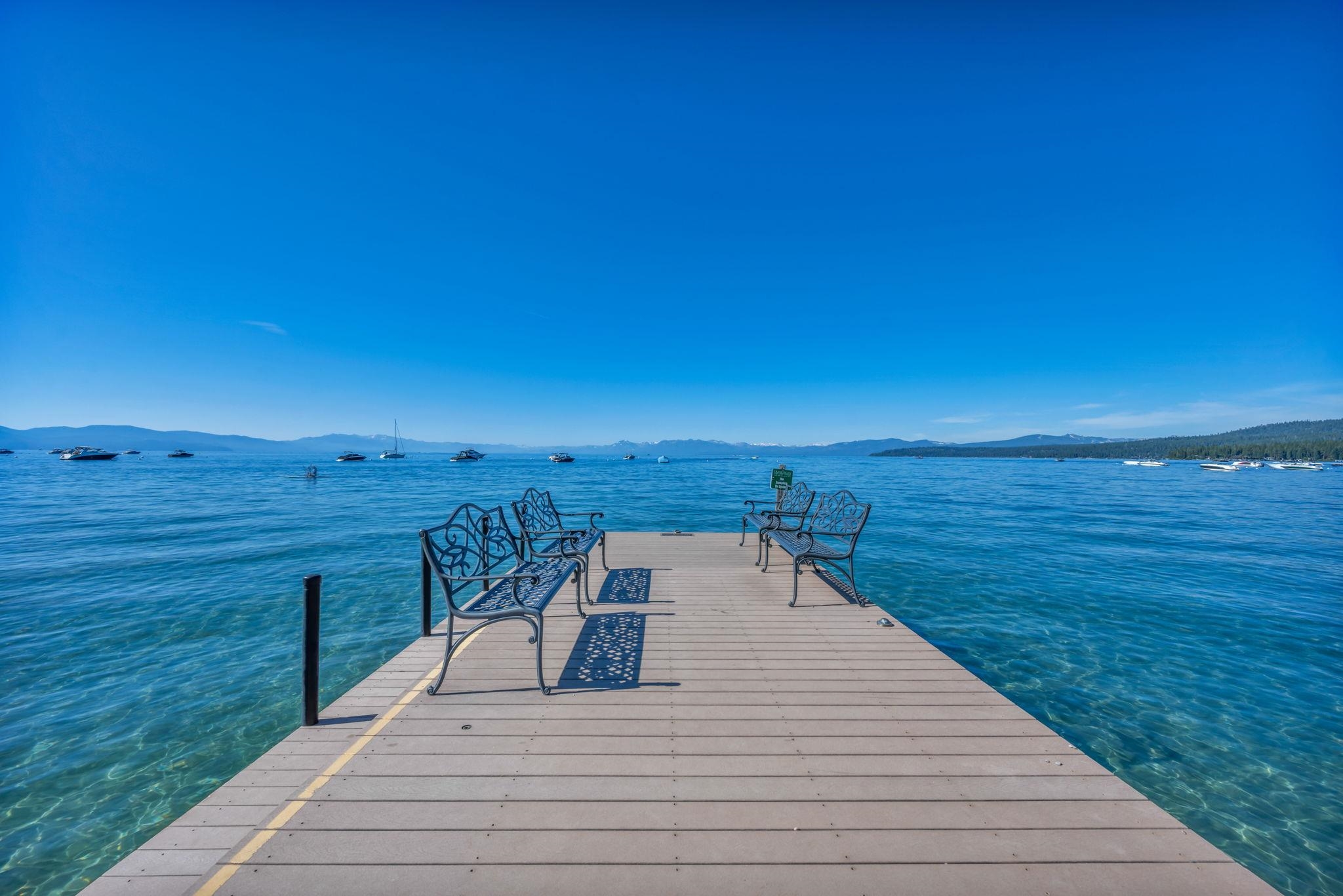 Dock with metal benches extends into a blue lake, with boats and distant mountains.
